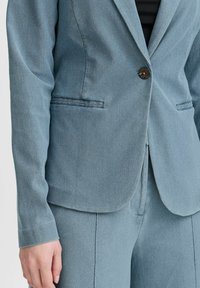 Úzký blazer v světle modré barvě z texturované látky, s jedním knoflíkem, dvěma předními kapsami a klopovým límcem.