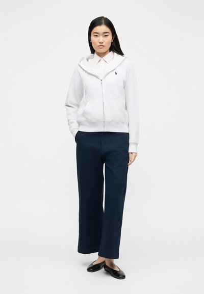 Νέα γυναίκα που στέκεται με τα χέρια στις τσέπες μπλε ναυτικού χρώματος παντελονιού, φοράει λευκό φούτερ με φερμουάρ, ριγέ πουκάμισο με γιακά και μαύρα παπούτσια τύπου μπαλαρίνας.