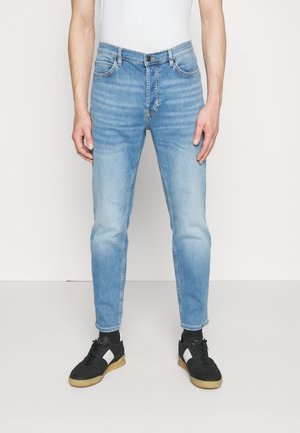 Jeans de mezclilla azul claro con un diseño ajustado, estilo de cinco bolsillos y sutiles detalles de desgastado en los muslos. Llevados con zapatillas deportivas negras.
