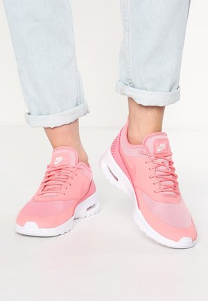 Stopy noszące podwinięte jasnoniebieskie dżinsy i jaskrawo różowe buty Nike Air Max Thea z białą podeszwą na białej podłodze.