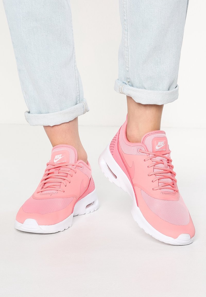 Des pieds portant un jean bleu clair retroussé et des baskets Nike Air Max Thea rose vif avec des semelles blanches sur un sol blanc.