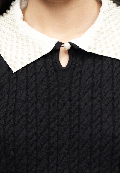 Gros plan sur un pull en maille noire avec un col Claudine blanc orné de petites perles et fermé par un gros bouton en forme de perle.