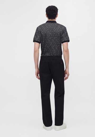 Homme debout, de dos, portant une chemise noire à manches courtes à motif, un pantalon noir et des baskets blanches sur un fond blanc uni.