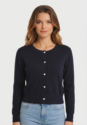 Donna con capelli castano chiaro che indossa un cardigan blu scuro aderente con bottoni a forma di cuore e jeans azzurri su uno sfondo semplice.