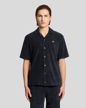 Junger Mann mit hellem Teint und zurückgekämmtem Haar trägt ein schwarzes, kurzärmeliges Hemd mit Knopfleiste und passende Shorts vor einem hellgrauen Hintergrund.