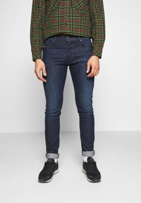 Mörkblå denimjeans med slim fit, uppvikta vid anklarna. I kombination med en grön och röd rutig skjorta och svarta sportskor.