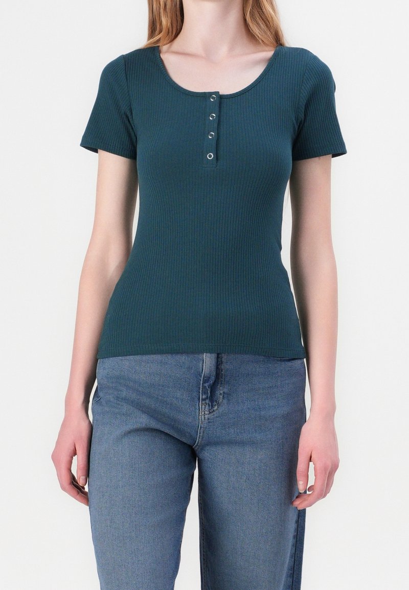 Chemise ajustée en maille côtelée, manches courtes et encolure dégagée, avec cinq boutons-pression métalliques sur le devant. Assortie à un jean bleu.