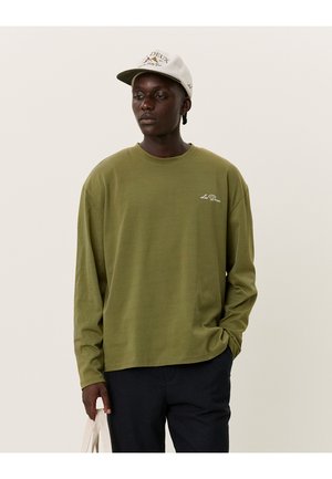 Cămașă cu mâneci lungi de culoare verde măsliniu, dintr-un material moale, cu guler rotund și un accent subtil cu logo. Se potrivește cu pantaloni închiși la culoare și o șapcă deschisă.