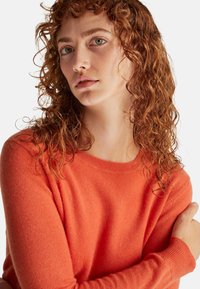 Młoda kobieta o kręconych rudych włosach, ubrana w pomarańczowy sweter, patrząca lekko na bok z neutralnym wyrazem twarzy, z założonymi rękami.
