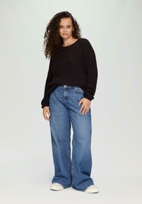 Schwarzer Strickpullover mit langen Ärmeln, kombiniert mit hellblauen weiten Jeans und weißen Leinen-Schuhen. Einfaches, lässiges Outfit vor einem neutralen Hintergrund.