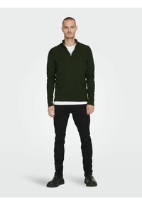 Dark green half-zip sweater made of textured knit fabric, paired with a white shirt and black fitted jeans. Black boots complete the outfit.