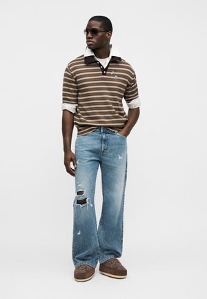 Man draagt een bruin gestreepte polo, gescheurde blauwe jeans met een grote scheur bij de knie, zonnebril en bruine schoenen, staat met één hand in zijn zak.