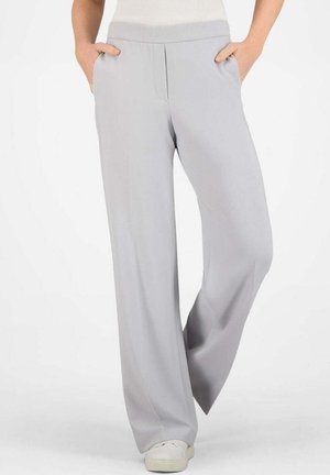 Person trägt hellgraue, weit geschnittene Hosen und weiße Turnschuhe, Hände in den Taschen, steht vor einem schlichten weißen Hintergrund.