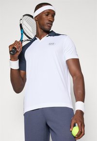 White polo shirt with navy accents, moisture-wicking fabric, featuring a logo. Holding a tennis racket and ball, with a white headband and wristband.