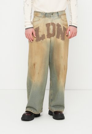 Jeans largos de ganga com coloração desbotada em castanho e verde, apresentando uma grande estampagem "LDN" na frente e desgaste subtil no tecido.