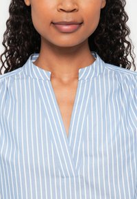 Lyseblå skjorte med krave og lodrette hvide striber, med en v-halsudskæring og diskret plissé nær skuldrene. Lavet af bomuldsmateriale.