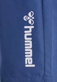 Blauer Stoff mit weißem "hummel" Markenlogo und stilisiertem Bienen-Symbol, vertikal auf der Oberfläche gedruckt.