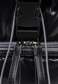 Bolso negro brillante con un broche negro, cremallera gris con tiradores metálicos y correas texturizadas con acentos de cuero sintético.