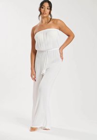 Mono blanco sin tirantes con textura plisada, parte superior elástica y cintura con cordón. Piernas de corte amplio que llegan hasta el suelo.