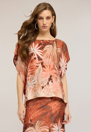 Kobieta o długich falowanych włosach, nosząca duże kolczyki-koła oraz zestaw luźnych ubrań: bluzkę i spódnicę z pomarańczowo-beżowymi tropikalnymi wzorami liści.