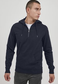 Mornarska zip-up kapuca z žepom spredaj, kapuco na vrvico, rebrastimi manšetami in teksturno poudarjenjem na robu. Mehka tkanina.