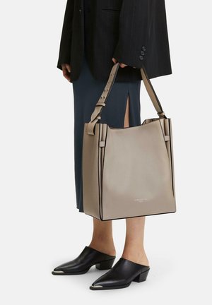 Une personne portant des mules noires et une jupe marine à fente tient un grand sac fourre-tout beige avec des anses larges et une bordure noire.