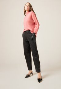 Mujer con suéter rosa, pantalones negros con tachuelas, guantes de cuero negros y tacones negros puntiagudos, posando frente a un fondo beige liso.