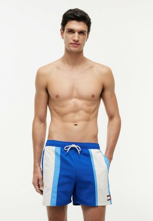 Young man standing with hands in pockets wearing blue and white striped swim shorts against a plain background.