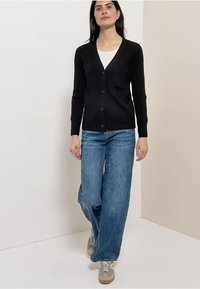 Schwarzer Cardigan mit V-Ausschnitt, Rippenstruktur und fünf Knöpfen, kombiniert mit hellblauen Jeans mit weitem Bein und beigen Sneakers.