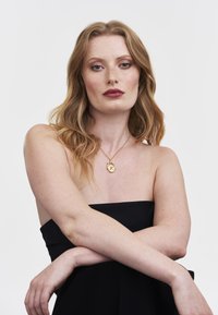 Gold pendant necklace with a circular charm and initials, worn over a black strapless garment. Model with medium-length wavy hair.