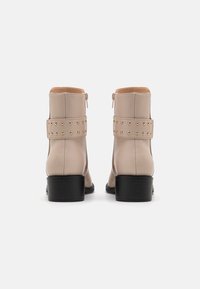 Beige kožené kotníkové boty se zlatými cvočky na popruhu, zaoblenou špičkou a černým robustním podpatkem. Zapínání na zip na zadní straně.