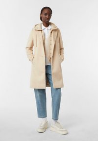 Cappotto lungo beige con cappuccio, tasche frontali e bottoni nascosti, indossato sopra un maglione leggero e una camicia; abbinato a jeans azzurri chiari e sneakers.