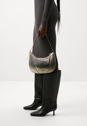 Woman in a ribbed gray dress and black knee-high boots holding a textured metallic gold handbag by its thin strap.