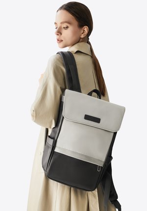 Zaino grigio e nero con chiusura a patta, dotato di una tasca frontale, spallacci e una maniglia superiore. Materiale liscio, design minimalista.
