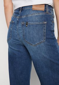 Tumman siniset farkut korkealla vyötäröllä, hienovaraisella haalistumisella, kaarevilla takataskuilla ja tekonahkatarra vyötäröllä bränditiedoilla.