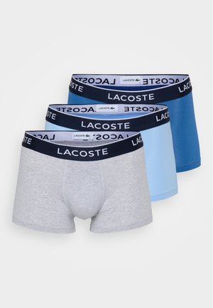 Pack of three men’s boxer shorts in grey, light blue, and dark blue. Each features an elastic waistband with "LACOSTE" logo. Cotton material.