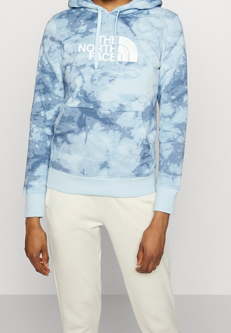 Personne portant un sweat à capuche tie-dye bleu clair avec le logo blanc "The North Face" et un pantalon couleur crème, debout, les bras détendus le long du corps.