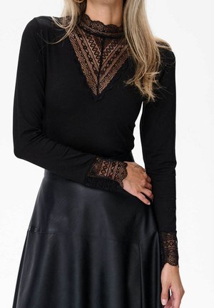 Femme portant un haut noir à manches longues avec un col en V en dentelle transparente et des détails aux poignets, associé à une jupe en cuir noire sur un fond uni.