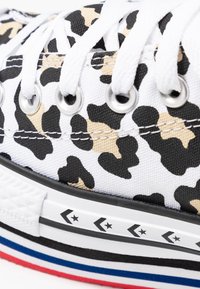 Primo piano di sneaker in tela bianca con stampa leopardata nera e beige, lacci bianchi, occhielli argentati e suola in gomma con strisce nere, blu e rosse.