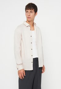 Young man looking right wearing beige buttoned shirt over white tank top and dark gray wide-leg trousers against plain white background