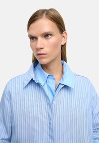 Jasnoniebieska koszula z długim rękawem w paski, z kontrastowym, jednolitym niebieskim kołnierzykiem, wykonana z miękkiej bawełny. Subtelny połysk podkreśla teksturę.