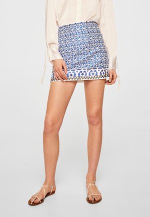 Kobieta ubrana w niebieską i białą mini spódniczkę w wzory, beżową bluzkę z długim rękawem oraz beżowe płaskie sandały na paski, stojąca na tle jednolitego tła.