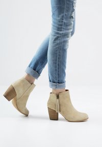 Beige suède enkellaarsjes met een opvallende blokhak en ritssluiting, voorzien van een franje-detail en een ribbelige houtachtige hak.