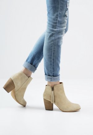 Beige semišové kotníkové boty s výrazným blokovým podpatkem a zapínáním na zip, zdobené detailem s třásněmi a žebrovaným podpatkem podobným dřevu.