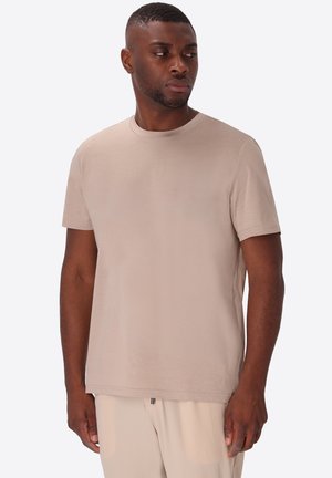 Uomo che indossa una maglietta beige a maniche corte e pantaloni abbinati, in piedi e guarda leggermente verso sinistra su uno sfondo semplice.