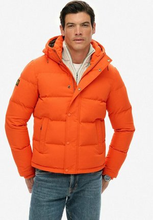Lys orange puffjakke med hætte, der har en quiltet tekstur, frontlommer og trykknaplukninger, lagdelt over en lys sweater.