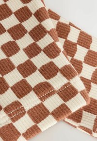 Close-up of ribbed fabric with brown and beige irregular checkerboard pattern and visible stitching on folded edges.
