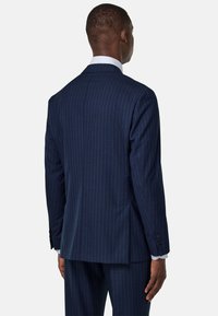 Veste de costume bleu marine à rayures fines avec une coupe ajustée, fente simple et boutons aux poignets. Tissu lisse avec un léger éclat. Chemise habillée blanche visible.