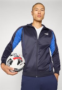 Marineblå treningsjakke med blå detaljer, glidelås foran og PUMA-logo, båret av en person som holder en hvit fotball med svarte og røde detaljer.