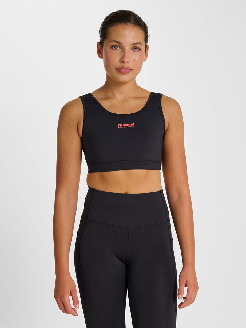 Frau steht, trägt ein schwarzes Sport-Crop-Top mit rotem „hummel performance“ Logo und hochtaillierte schwarze Leggings vor weißem Hintergrund.